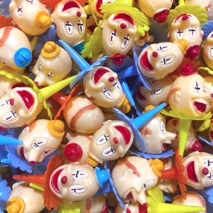 May include: A collection of vintage clown-shaped food picks. The plastic clown heads are cream-colored with red painted features, including a large red smile. The picks have colorful accents, including blue, yellow, and orange.