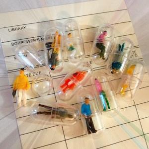May include: A collection of clear plastic capsules containing miniature figurines of people in various poses. The capsules are arranged on a lined paper surface.