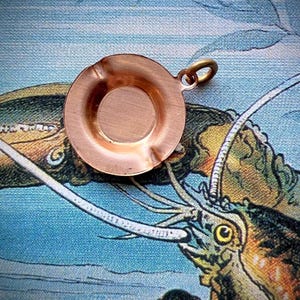 May include: A small, round, copper-colored ashtray with a circular indentation in the center and a small loop for hanging. The ashtray is set against a backdrop of a blue and green illustration of a lobster.