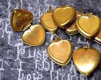 May include: A group of eight gold heart-shaped locket pendants with hinges. The lockets are arranged in a cluster on a gray fabric background.