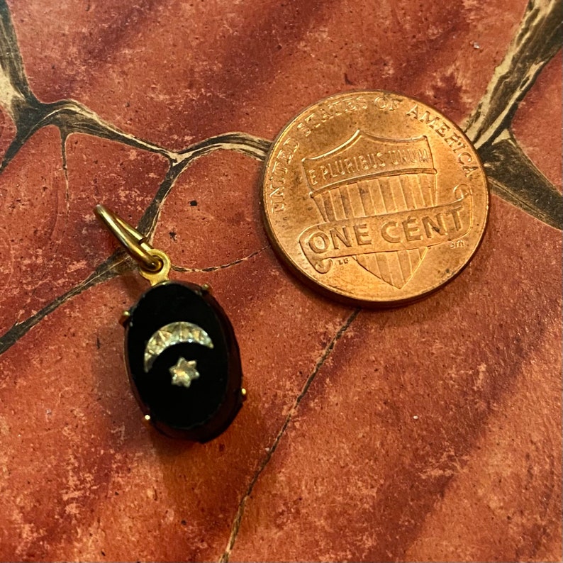 May include: A gold-toned pendant with a black oval center, featuring a crescent moon and star design. A U.S. one-cent coin is next to the pendant for size comparison. The pendant has a small loop at the top for a chain.
