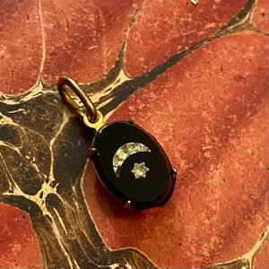 May include: A gold-toned pendant with a black, oval-shaped center. The center features a crescent moon and a star, both embellished with small, sparkling stones. The pendant has a loop for a chain.