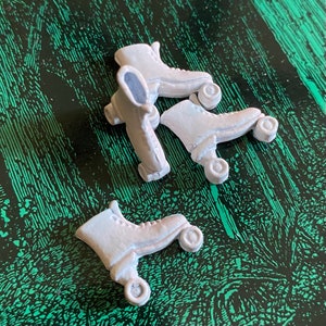 May include: Three white roller skates with black wheels. The skates are arranged in a pile, with the wheels facing up.