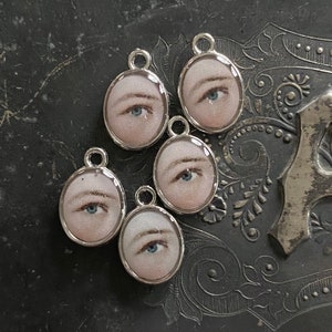 Puede incluir: Cinco dijes de plata con fondo blanco y un ojo pintado con iris azul. Los dijes son de forma ovalada y tienen un bucle en la parte superior para sujetarlos a un collar o pulsera.