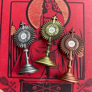 May include: Three gold, silver, and copper religious ornaments with a sunburst design and the letters "JHS" in the center. The ornaments are on stands and have a cross on top.