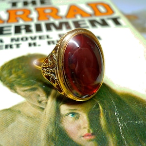 May include: A gold ring featuring a large, oval-shaped, dark red gemstone. The ring is resting on a book with the title "The Arrad Experiment" printed on the cover.