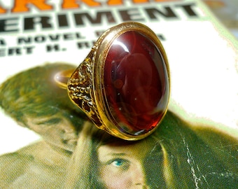 May include: A gold ring with a large, oval-shaped, dark red gemstone. The ring is sitting on top of a book with the title "The Arrad Experiment" printed on the cover.