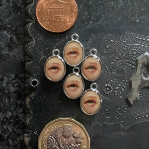 May include: Six silver charms with images of eyes on a dark background. The charms are arranged in a circle around a US penny and a British pound coin.