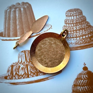 May include: A copper-toned pendant with a textured surface and a small spoon-shaped charm. The pendant has a loop at the top for hanging. The background features illustrations of desserts in shades of brown.