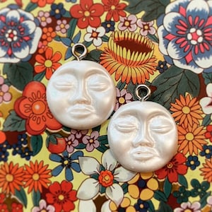 May include: Two white ceramic moon-shaped charms with closed eyes and a slightly parted mouth. The charms have a shiny, pearlescent finish and are hanging from silver loops.