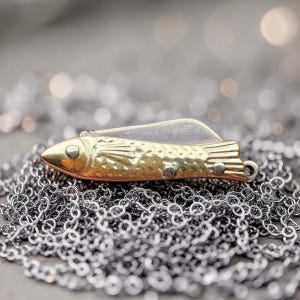 Könnte beinhalten: Ein goldfarbener Fischanhänger mit strukturiertem Körper und silberfarbener Klinge, der auf einer silbernen Kette liegt. Der Fischanhänger hat am Ende eine kleine Schlaufe zum Befestigen an einer Halskette. Der Hintergrund ist verschwommen, mit Bokeh-Lichtern.