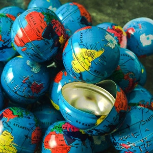 May include: A collection of vintage globe-shaped trinket boxes with various colors and designs. The globes feature maps of the world with continents and oceans labeled in different languages. Some of the globes are open, revealing a silver interior.