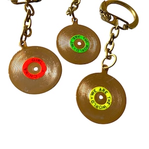 May include: Three keychains with brown record-shaped pendants. One pendant has a red center with "CHINA GIRL" text, while the others have green and yellow centers with "WE ARE THE WORLD" text. The keychains have gold-colored chains and clasps.