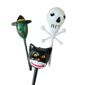 以下が含まれることがあります： ハロウィンをテーマにした装飾が3つ。緑色の魔女が黒い帽子をかぶり、黒い猫が黄色い目をしている。白い骸骨は骨が交差している。