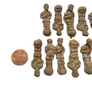 May include: A collection of small, weathered, gray-brown figurines, each depicting a standing human figure. A U.S. one-cent coin is included for scale. The figurines appear to be antique or vintage, possibly made of stone or a similar material.