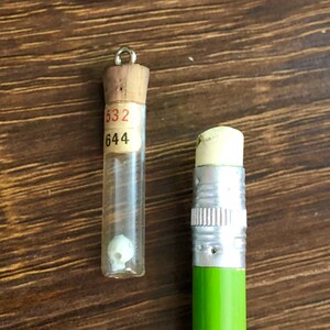 May include: A small glass vial with a cork stopper and a label with the numbers "532" and "644" on it. The vial contains a small white object. A green pencil with a silver eraser is next to the vial.
