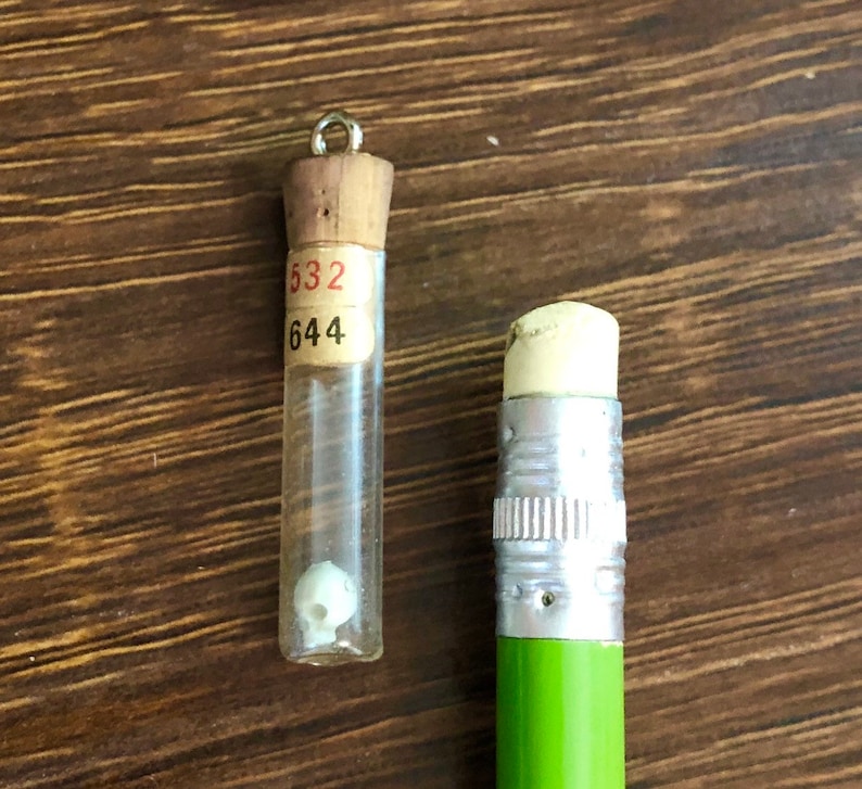 May include: A small glass vial with a cork stopper and a label with the numbers "532" and "644" on it. The vial contains a small white object. A green pencil with a silver eraser is next to the vial.