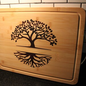 Può includere: Un tagliere in bambù marrone con un design ad albero della vita inciso sulla superficie. Il tagliere ha due manici argentati sui lati.