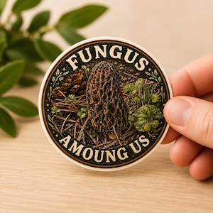 May include: A round sticker featuring a detailed illustration of a morel mushroom, pine cone, and greenery. The sticker has the words "FUNGUS AMONG US" in a circular design around the central image. The sticker is held by a hand.