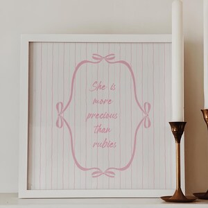 Könnte beinhalten: Ein weißer gerahmter Druck mit rosa Streifen und einem rosa kursiv geschriebenen Zitat, das "She is more precious than rubies" innerhalb eines rosa verzierten Rahmens lautet. Der Druck befindet sich auf einem weißen Regal mit zwei weißen Kerzen in goldenen Kerzenhaltern.