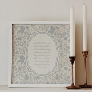 May include: A framed print with a floral design and the text "The Lord bless you and keep you...and give you peace." Two white candles stand in brass candle holders. The print is in a white frame.