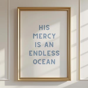 May include: A framed print with the words "HIS MERCY IS AN ENDLESS OCEAN" in light blue lettering. The print is in a gold frame and hangs on a white wall with molding.