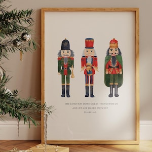 May include: A framed print featuring three watercolor nutcracker soldiers in red, green, and black uniforms. The print includes the text "THE LORD HAS DONE GREAT THINGS FOR US AND WE ARE FILLED WITH JOY" and is displayed near a Christmas tree.