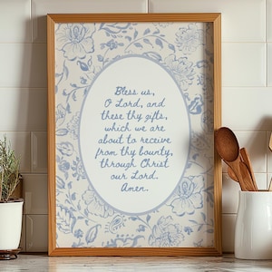 Op de afbeelding: Een ingelijste print met een licht houten frame. De print heeft een bloemenpatroon in lichtblauw en wit. Een ovaal in het midden bevat een gebed in blauwe tekst. De print wordt weergegeven op een aanrecht met keukenartikelen.