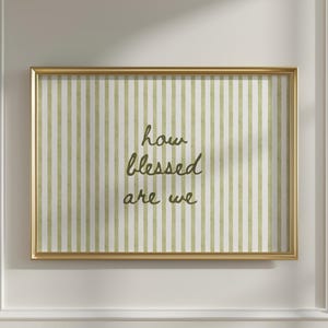 May include: A framed print with a gold border, featuring a striped background in white and olive green. The text "how blessed are we" is written in a cursive font. The artwork is displayed on a white wall.