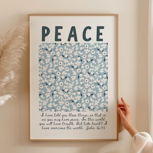 May include: A framed art print with the word "PEACE" in dark blue letters. The print features a light blue floral pattern and a quote from John 16:33. The frame is a light brown color.