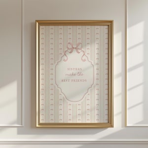May include: A framed print with a gold-colored frame. The print features a light pink and cream-colored background with vertical stripes and bow accents. The text "sisters make the best friends" is displayed in a decorative oval shape.