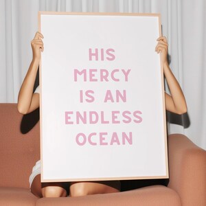May include: A framed poster with the text "HIS MERCY IS AN ENDLESS OCEAN" in pink lettering. The poster is held up in front of a person sitting on a peach-colored sofa. White curtains are in the background.