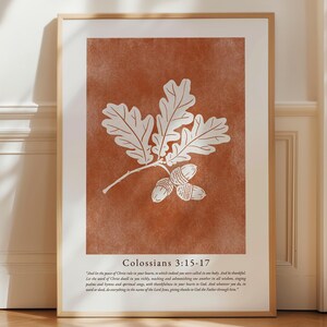 May include: A framed print featuring a white oak leaf and acorn illustration on a textured, rust-colored background. The print includes the text "Colossians 3:15-17" and a Bible verse. The frame is a light brown color.
