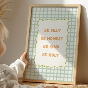 Op de afbeelding: Een ingelijste print met de woorden "BE SILLY, BE HONEST, BE KIND, BE HOLY" in oranje tekst. De print heeft een lichtgroene geruite achtergrond en een licht houten frame. Een kind houdt de ingelijste print vast.