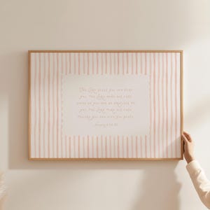 May include: A framed print with a light wood frame. The print features a pink and white striped background with a central rectangular area containing handwritten text. The text is a religious quote from Numbers 6:24-26.