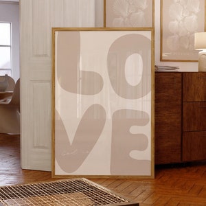 May include: A framed print with a beige background and the word "LOVE" written in large, stylized, beige letters. The letters are rounded and have a slightly textured appearance. The word "Good is" is written in a smaller, cursive font at the bottom of the print.