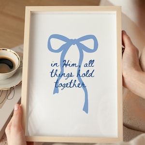 May include: A framed print with a light wood frame. The print features a blue bow illustration and the words "in Him, all things hold together" in a matching blue script. A cup of coffee and a book are in the background.