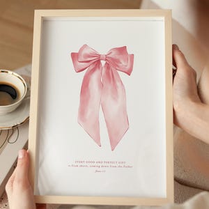 Puede incluir: Una impresión enmarcada con una ilustración de acuarela de un lazo rosa con largas cintas. La obra de arte incluye el texto "EVERY GOOD AND PERFECT GIFT is from above, coming down from the Father." Una taza de café y un libro están en el fondo.