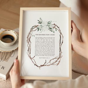 Puede incluir: Impresión enmarcada con el texto "THE RESURRECTION STORY" y un diseño de corona de flores y ramitas. El texto es de Mateo 28:1-7. El marco es de madera clara.