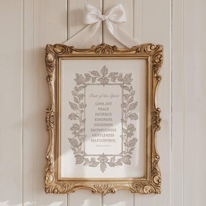 May include: A framed print with the text "Fruit of the Spirit" and a list of virtues, in a gold ornate frame. The print is hung with a white ribbon and bow against a white wood-paneled wall.