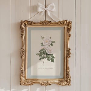May include: Ornate gold framed print of white roses with green leaves, hanging from a white bow. The print has a light blue border and a quote at the bottom. The frame has detailed carvings and is mounted on a white wooden wall.