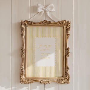 May include: A gold ornate picture frame with a yellow and white striped background and the text "You are my sunshine, my only sunshine." A white bow and ribbon are attached to the top of the frame.