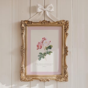 Op de afbeelding: Een ingelijste bloemenprint met roze en witte rozen, in een gouden, sierlijke lijst. De print heeft een roze passe-partout en wordt opgehangen met een witte satijnen strik. Het kunstwerk wordt tentoongesteld tegen een witte houten muur.