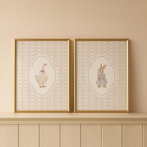 May include: Two framed prints featuring a goose and a rabbit. The goose wears a pink bow, and the rabbit is in a dress. The background is a striped pattern in cream and pink. The frames are gold.