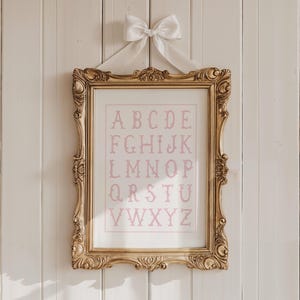 May include: A framed alphabet print in a gold ornate frame, hanging on a white wooden wall. The print features the alphabet in a soft pink color. A white bow is tied at the top of the frame, adding a decorative touch.