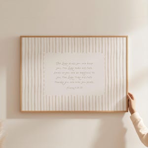 Op de afbeelding: Een ingelijste print met een lichtbeige en wit gestreepte achtergrond. De print bevat een handgeschreven script van een religieus vers in lichtbruin. De lijst is van lichtbruin hout.