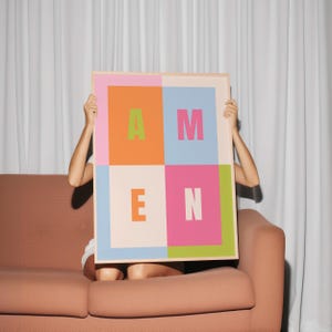 Könnte beinhalten: Ein gerahmter Kunstdruck mit dem Wort "AMEN" in großen, farbenfrohen Blockbuchstaben. Die Buchstaben sind in Grün, Pink, Orange und Weiß gehalten und vor einem hellen Hintergrund platziert. Das Kunstwerk wird von einer Person gehalten, die auf einem pfirsichfarbenen Sofa sitzt.