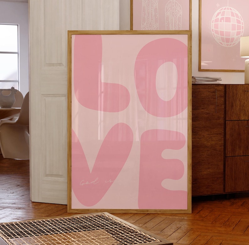 May include: A framed pink and white print with the word "LOVE" in large, stylized letters. The letters are outlined in a light pink color and the background is a gradient of pink and white. The text "Good is" is written in a smaller font at the bottom of the print.