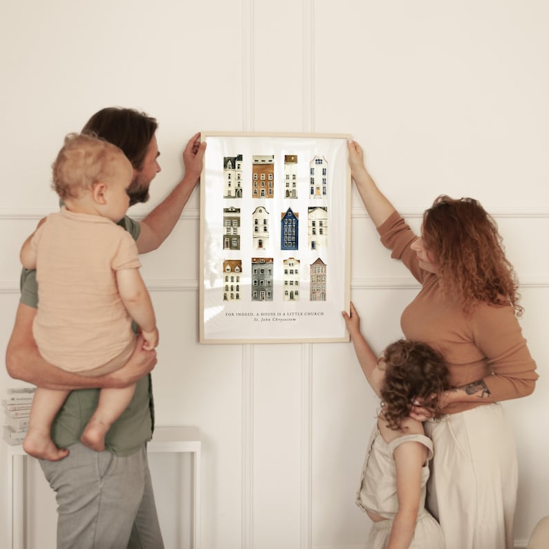 May include: A framed print featuring a collection of illustrated buildings in various colors. The print is being held by a family. The text at the bottom reads "FOR INHER, A HOUSE IS A LITTLE CHURCH by John O'Donohue."