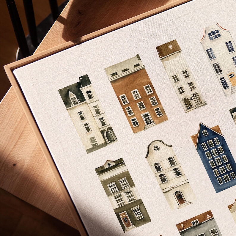 May include: Watercolor painting of various European-style buildings in shades of brown, white, and blue. The artwork is framed in a light brown wooden frame. The buildings have multiple windows and unique architectural details.
